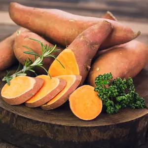 Raw sweet potatoes, whole and chopped up with other vegetables like rosemary and brocolli on a wooden chopping board.